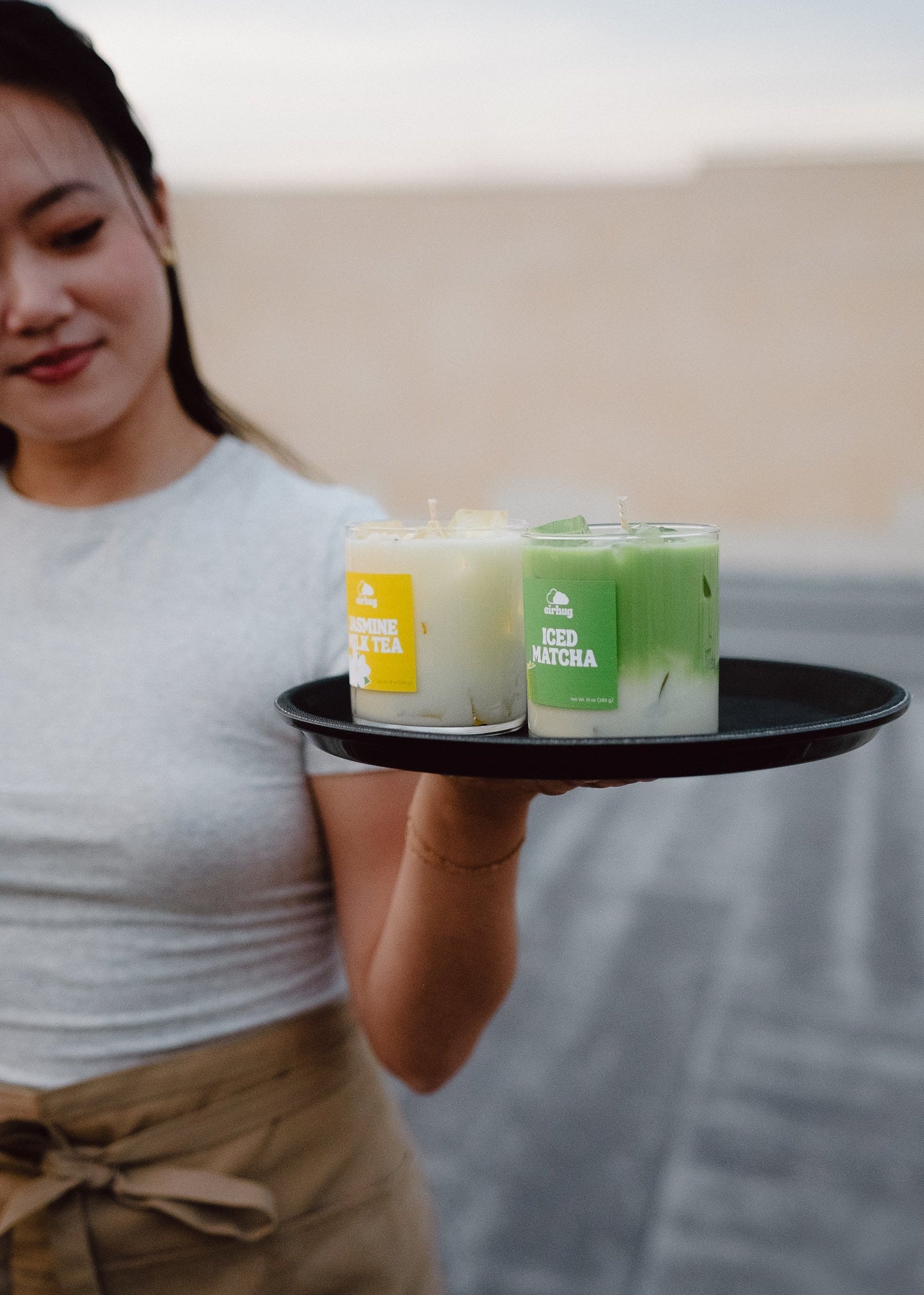 Person holding a tray with two candles, one labeled 'Iced Matcha' and the other 'Jasmine Milk Tea', against a blurred background.