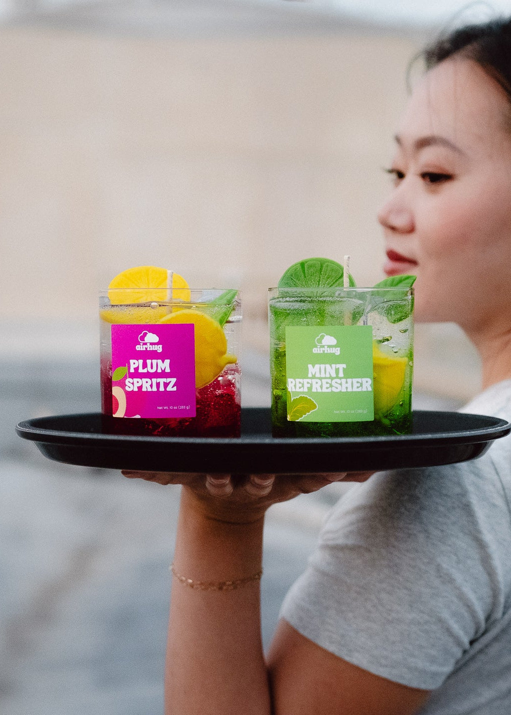 Person holding a tray with two cocktail inspired candles labeled 'Plum Spritz' and 'Mint Refresher'.