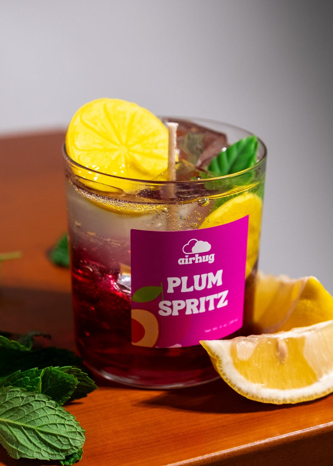 Glass of plum spritz candle with lemon slices and mint leaves on a wooden surface.