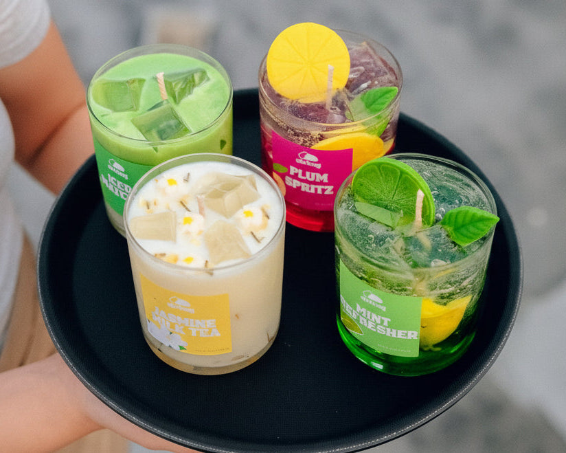 Four candles in glass jars with colorful labels on a black tray.