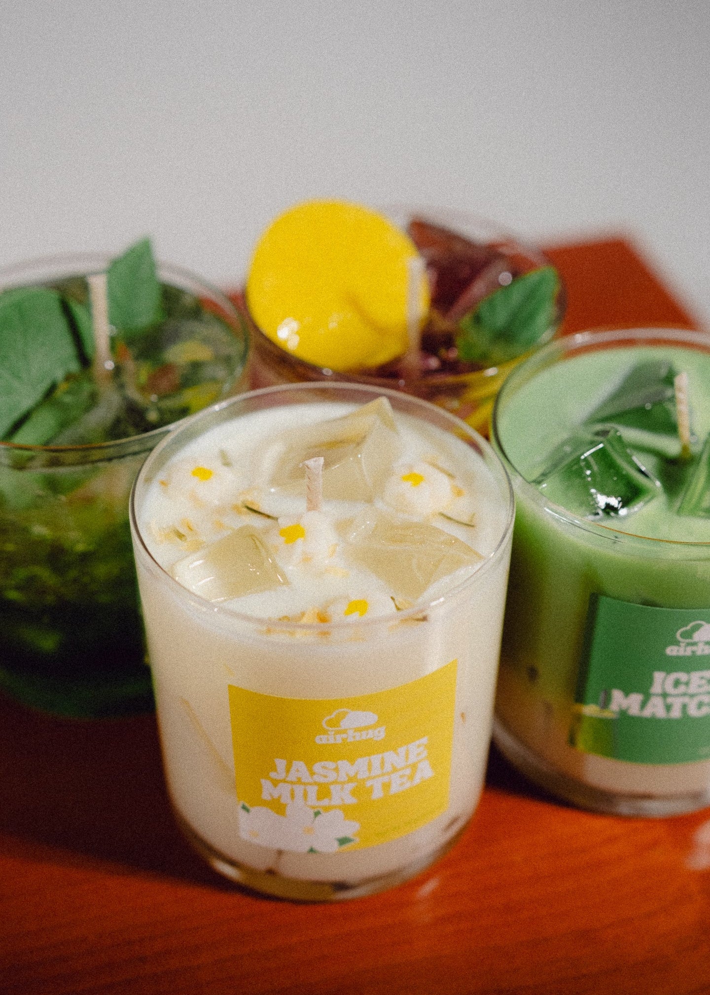 Four dessert candles, labeled 'Jasmine Milk Tea', 'Iced Matcha', and 2 hidden in the back on a wooden surface with a blurred background.